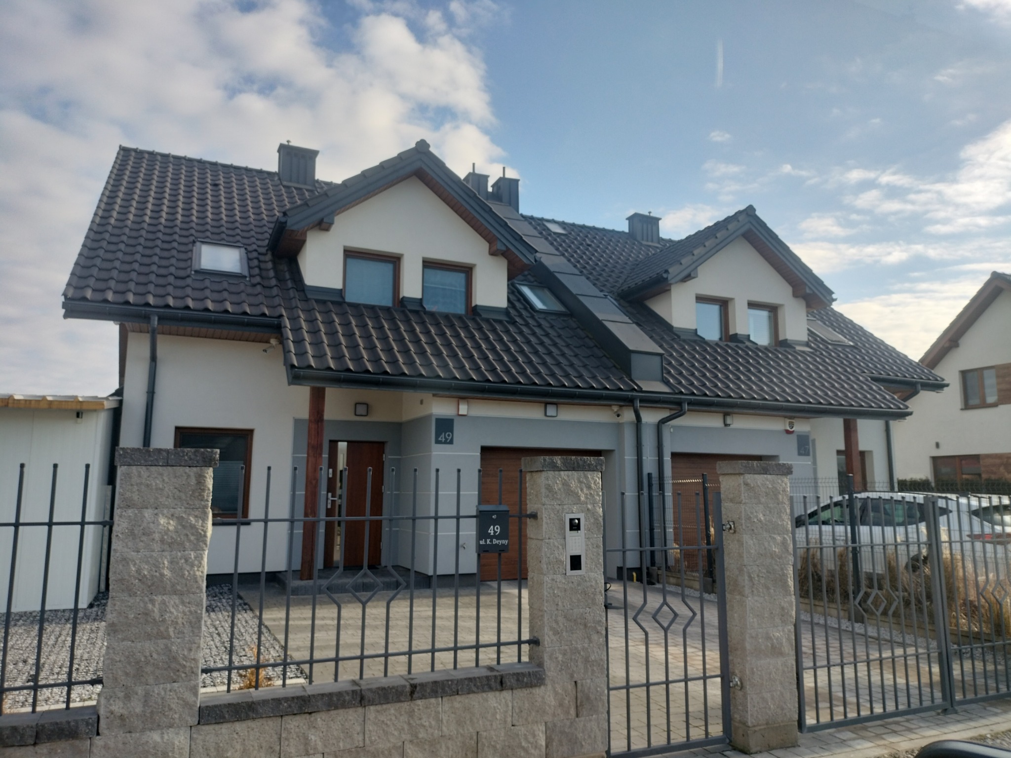 Dwukondygnacyjny dom z ciemną dachówką, oknami dachowymi i metalową obróbką blacharską, widziany zza ogrodzenia.