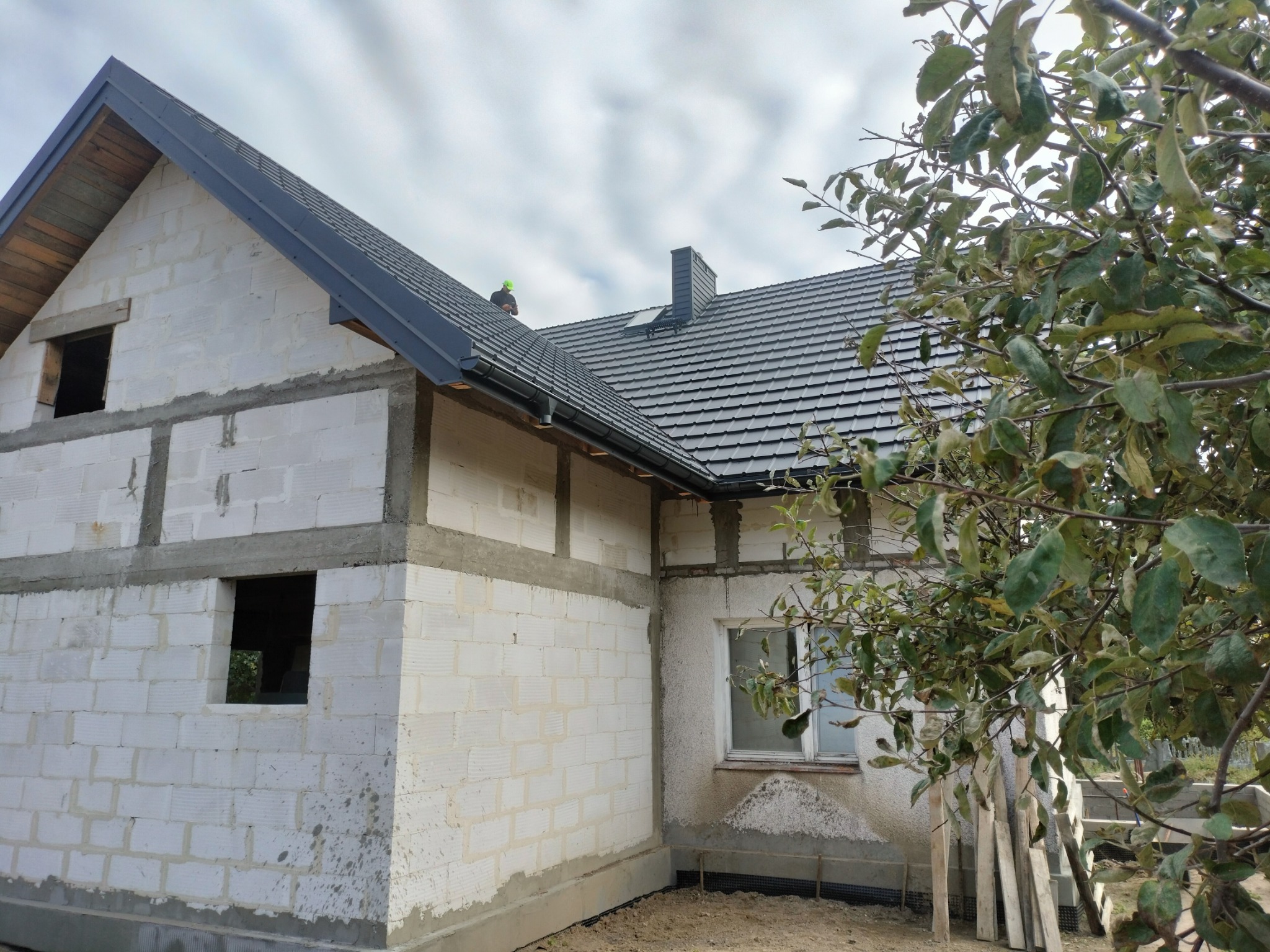 Surowy stan budynku z białego bloczka, widoczna czarna dachówka i obróbka blacharska, fragment rynny, okno i drzewo z liśćmi w kadrze.