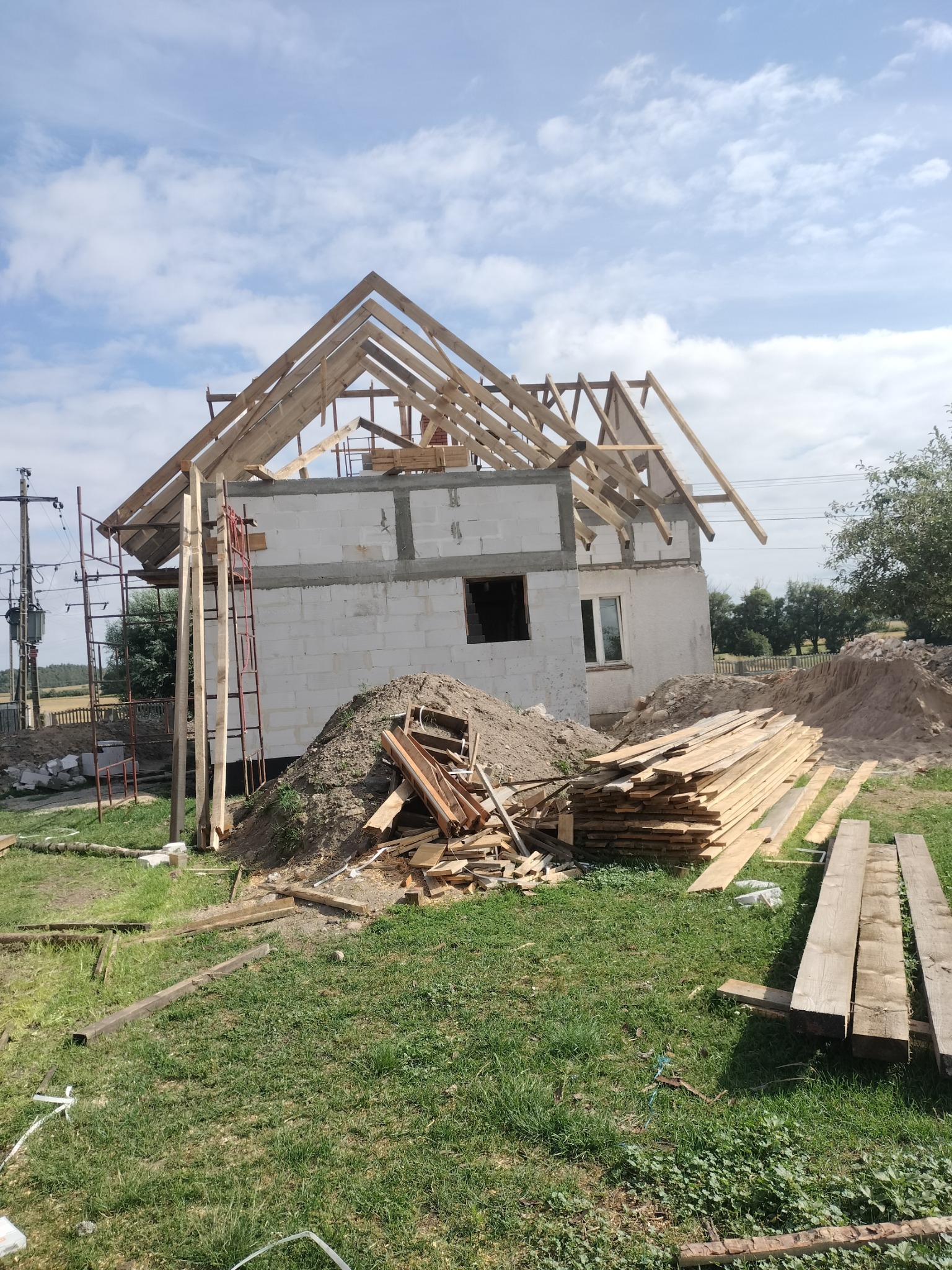 Surowy stan budowy domu jednorodzinnego: wzniesione ściany z bloczków, częściowo wykonana drewniana konstrukcja dachu, stosy desek i ziemi na trawiastym terenie.