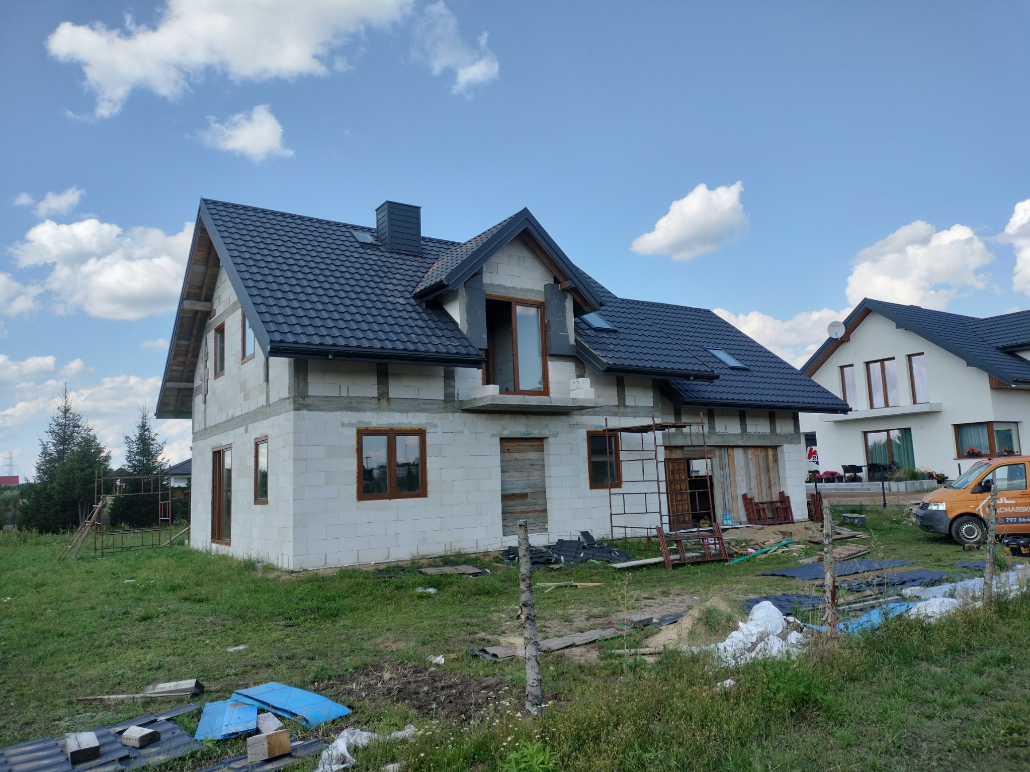 Dom w stanie surowym otwartym z ciemną dachówką, widoczna więźba dachowa, okna częściowo zamontowane, w tle błękitne niebo z chmurami.