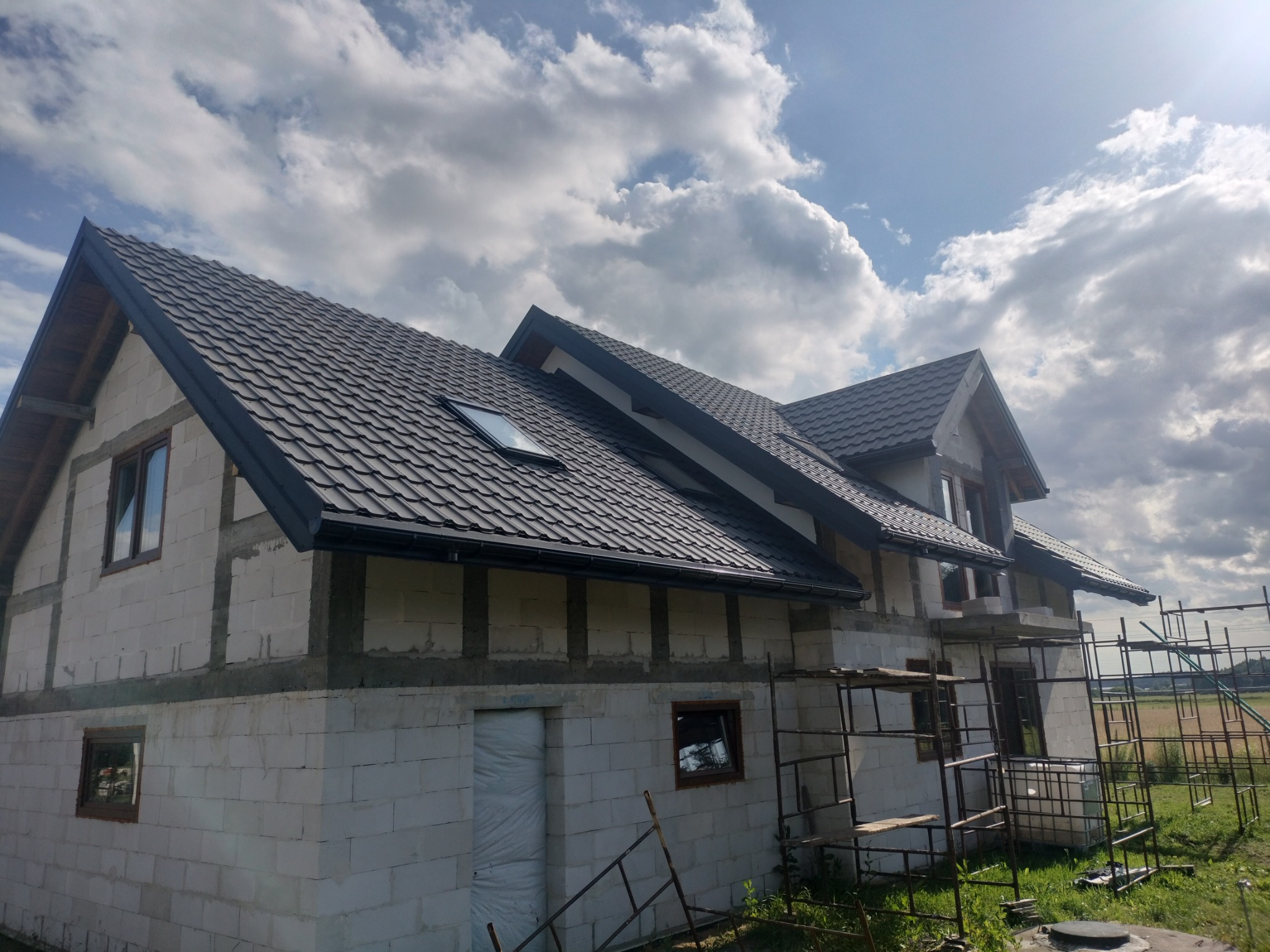 Dom w trakcie budowy z czarnym dachem z dachówki, widoczne rusztowania i ściany z białych bloczków, okna w drewnianych ramach.