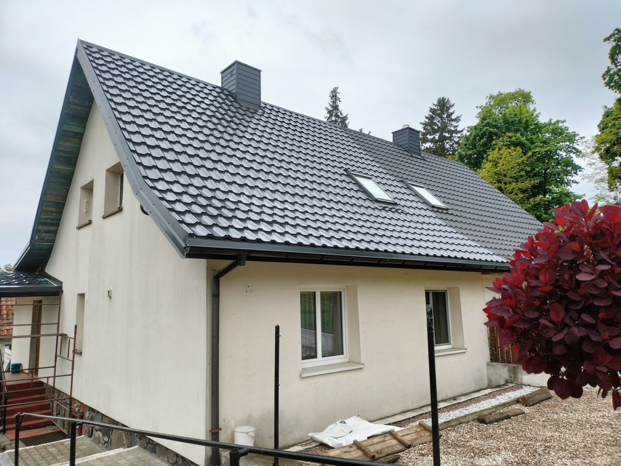 Dom jednorodzinny z nowym, ciemnoszarym dachem z dachówki, widoczne dwa kominy i okna dachowe, elewacja w kolorze kremowym, obok rośnie bordowy krzew.