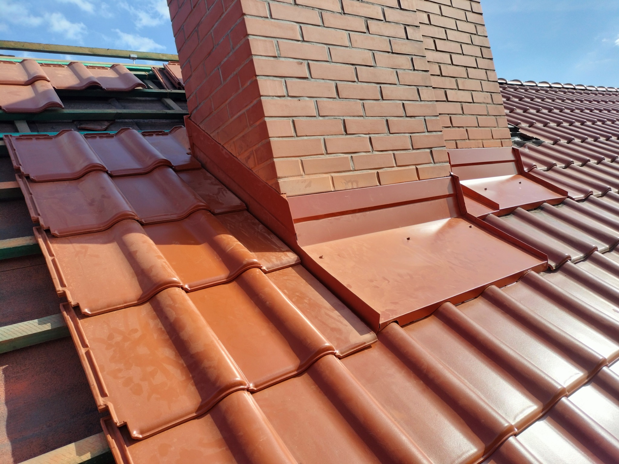 Ceglany komin z obróbką blacharską na dachu z czerwonej dachówki ceramicznej, widoczne elementy więźby dachowej i fragment nieba.