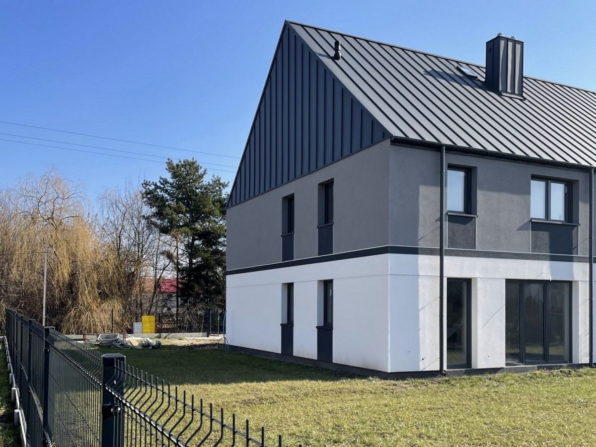 Nowoczesny dom dwukondygnacyjny z ciemnoszarą blachą na dachu i szarymi ścianami na górze, białe ściany na dole, widok z zewnątrz na tle zielonego trawnika i drzew bez liści.