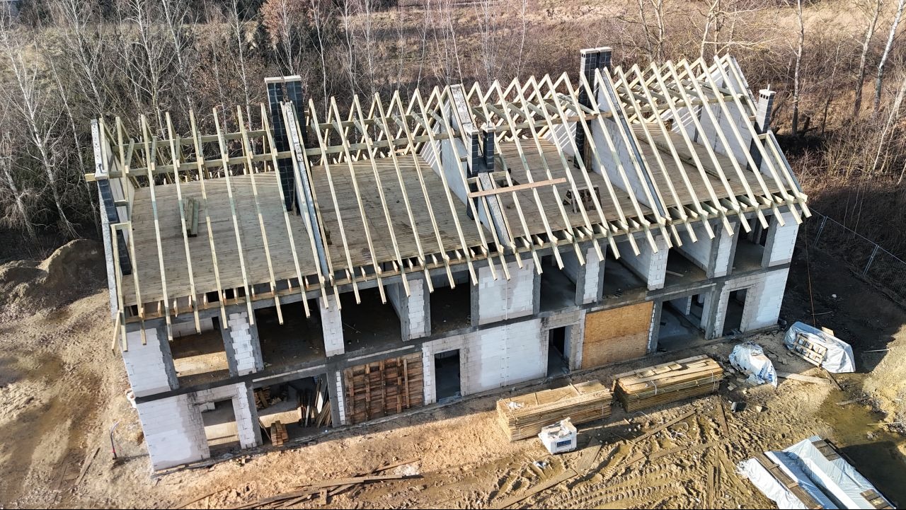 Surowa konstrukcja domu w trakcie budowy, widok z góry na dwukondygnacyjny budynek z pustaków z częściowo wykonaną drewnianą konstrukcją dachu i kominami, na ziemi leżą materiały budowlane.