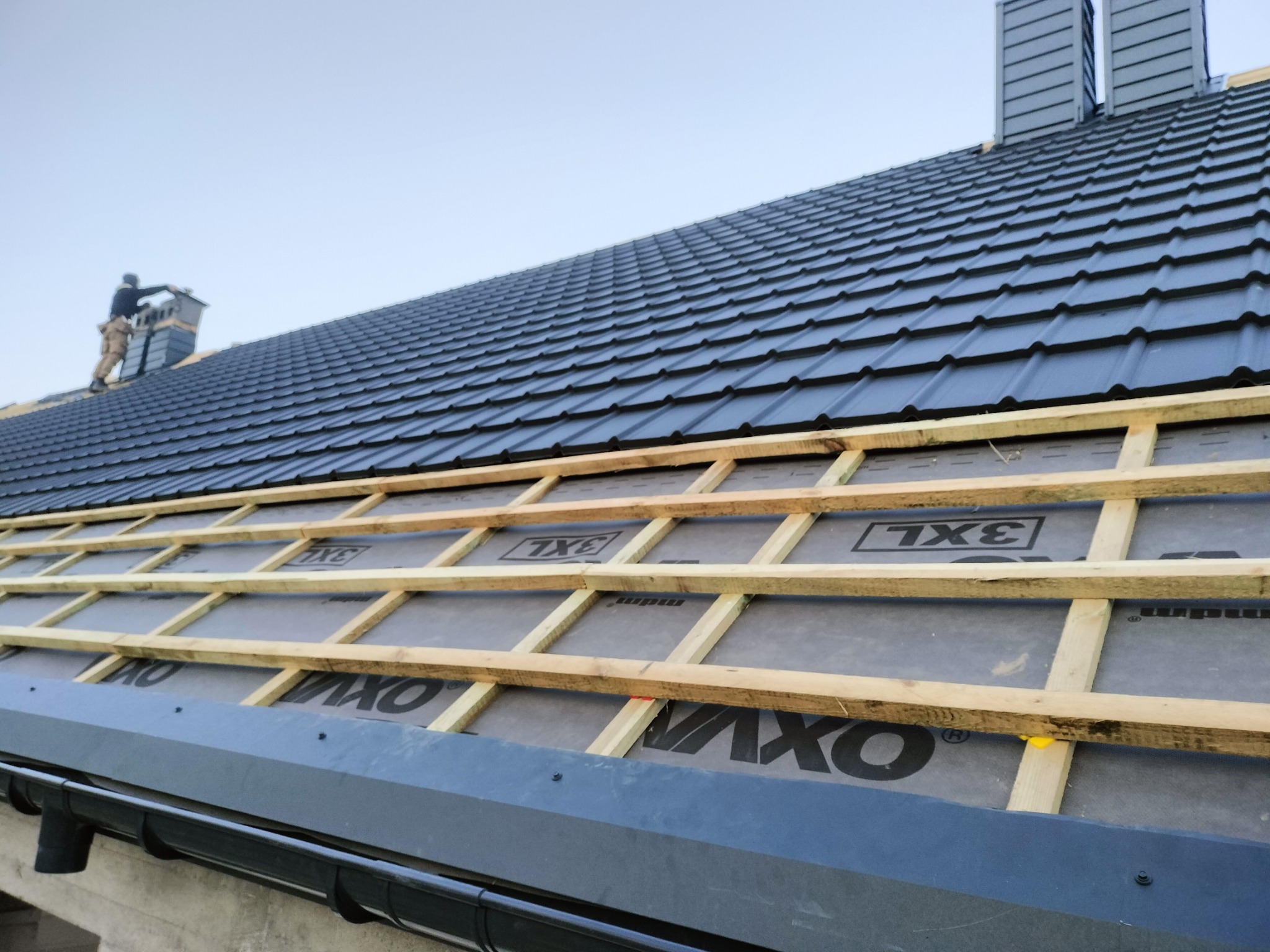 Montaż czarnej dachówki ceramicznej na łatach, widoczny dekarz przy kominie, orynnowanie w kolorze dachu.