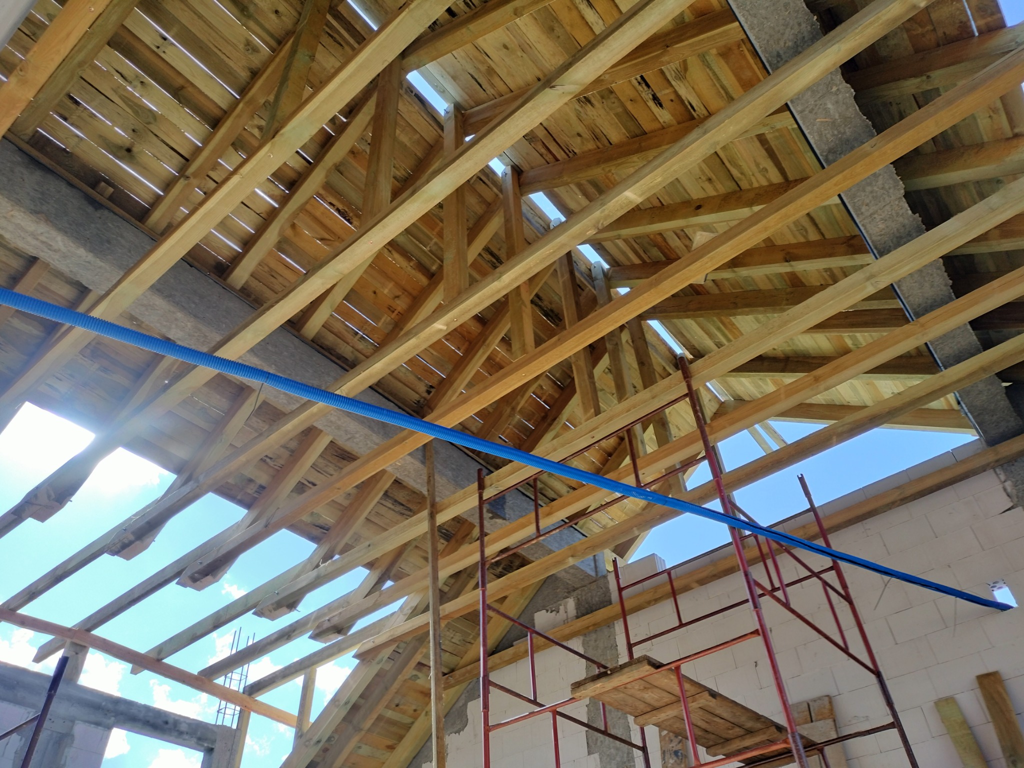 Wnętrze konstrukcji drewnianej dachu z widocznymi łatami, krokwiami i izolacją, na tle nieba widocznego przez otwory, z elementami rusztowania i niebieską rurą instalacyjną.