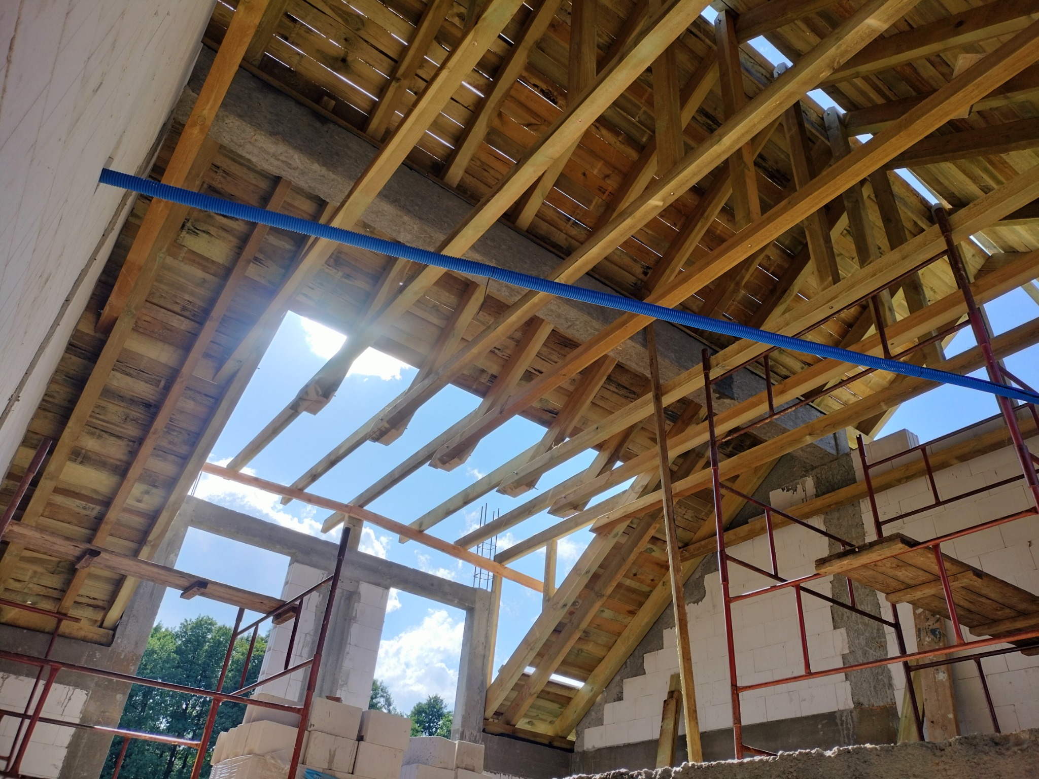 Wnętrze budynku w trakcie budowy z widoczną drewnianą konstrukcją dachu, rusztowaniem i niebieską rurą instalacyjną na tle nieba.