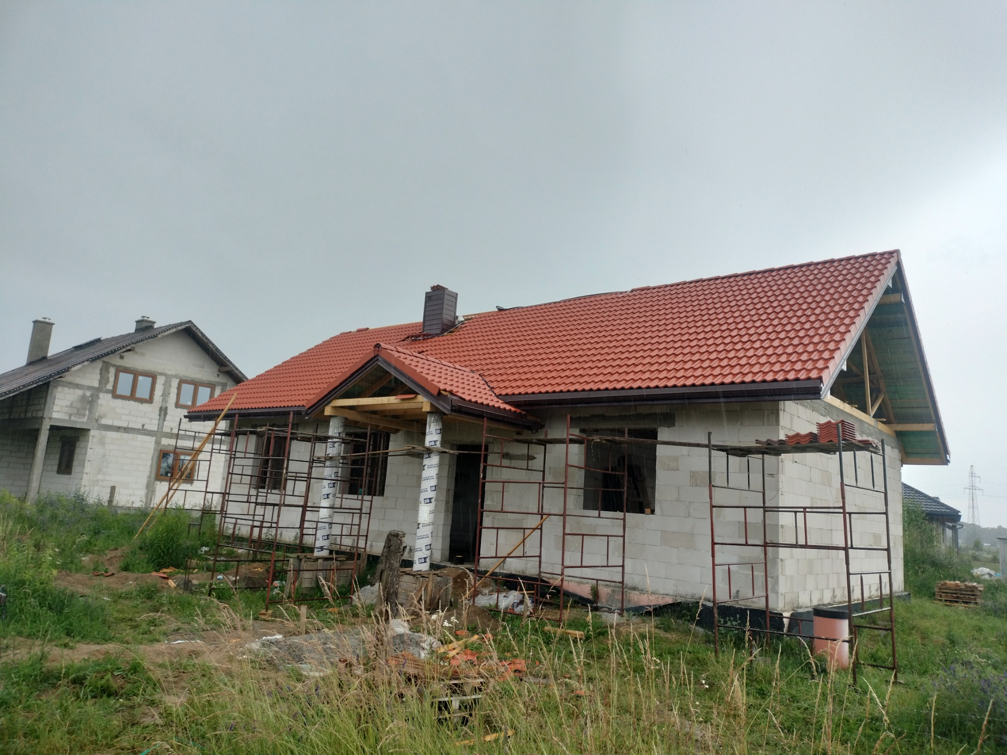Dom w trakcie budowy z częściowo położonym czerwonym dachem, widoczne rusztowania i elementy więźby dachowej, drugi dom w tle.
