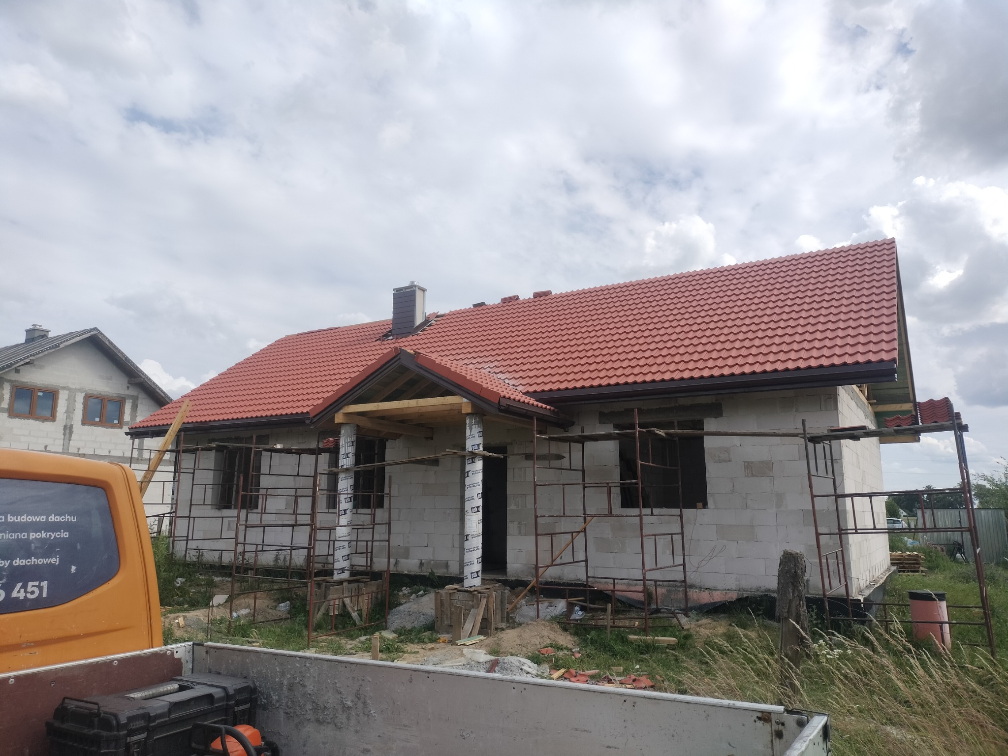 Dom w trakcie budowy z nowym, czerwonym dachem z dachówki ceramicznej, widoczne rusztowania i elementy konstrukcyjne z drewna.
