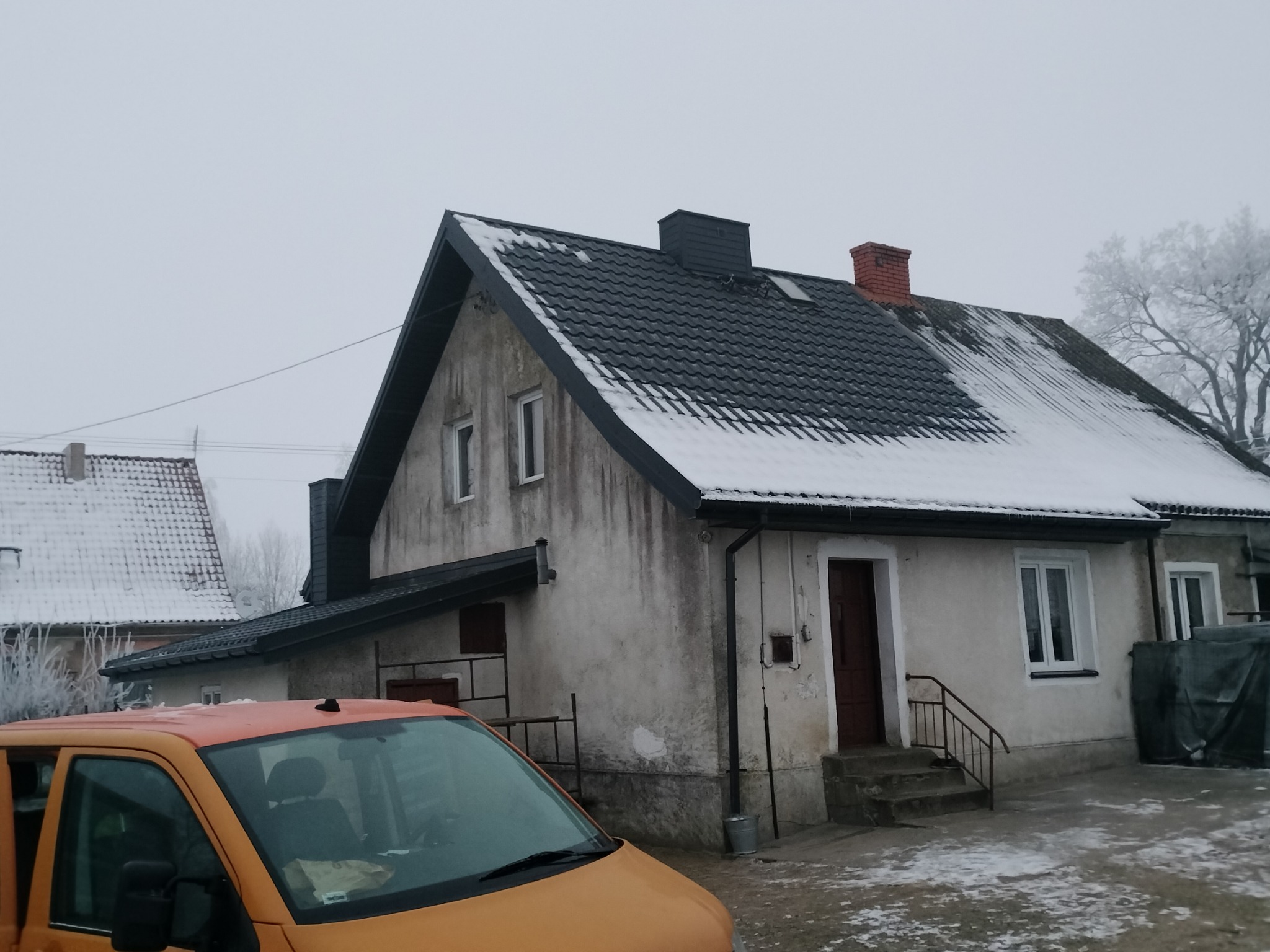 Dom jednorodzinny z ciemnoszarą dachówką, częściowo pokrytą śniegiem. Pomarańczowy samochód dostawczy na pierwszym planie. Widoczny komin z cegły i ośnieżone drzewa w tle.