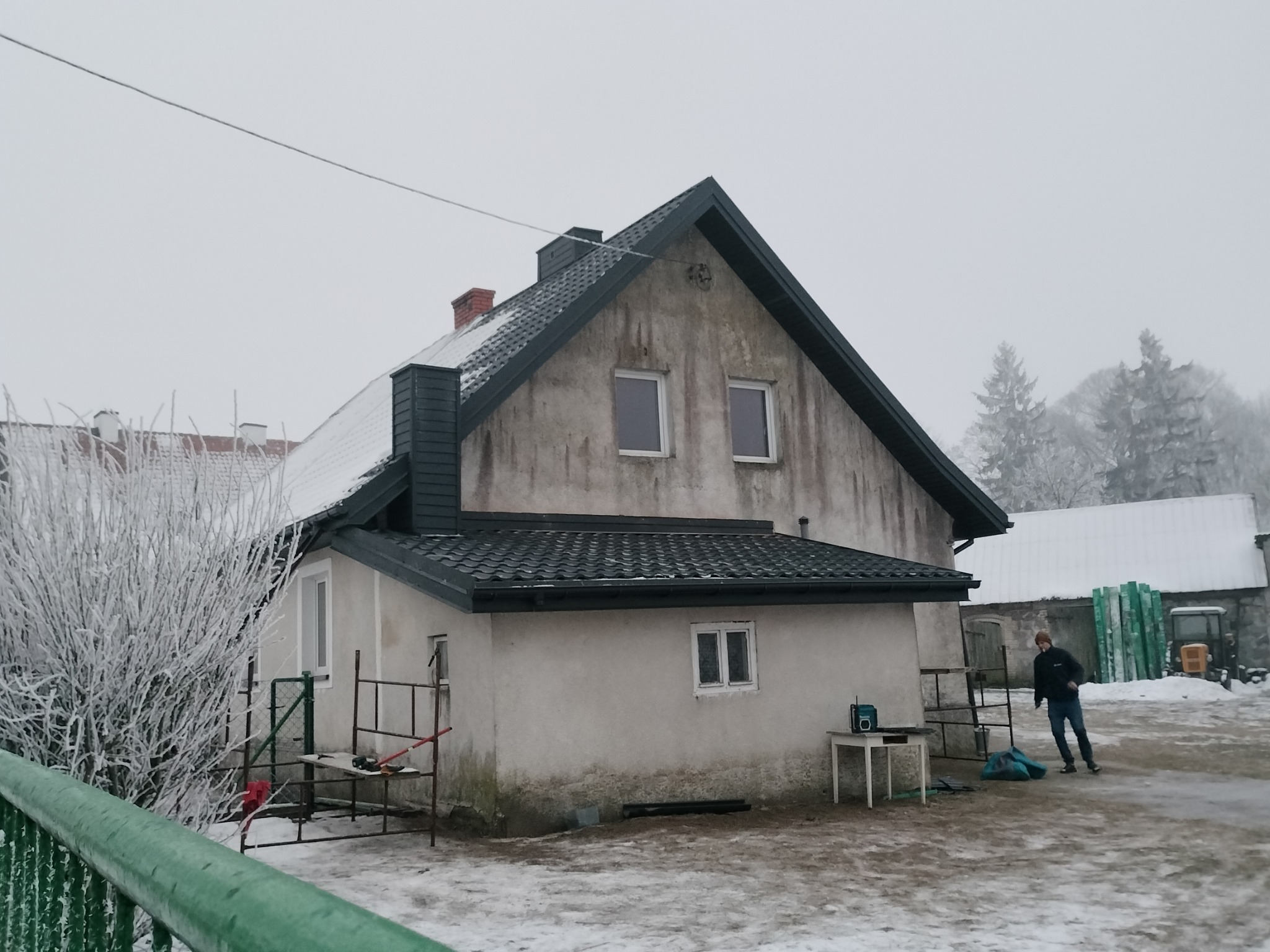 Dom jednorodzinny z nowym, ciemnym dachem z dachówki ceramicznej w zimowej scenerii, widoczne rusztowanie, narzędzia i osoba w oddali.