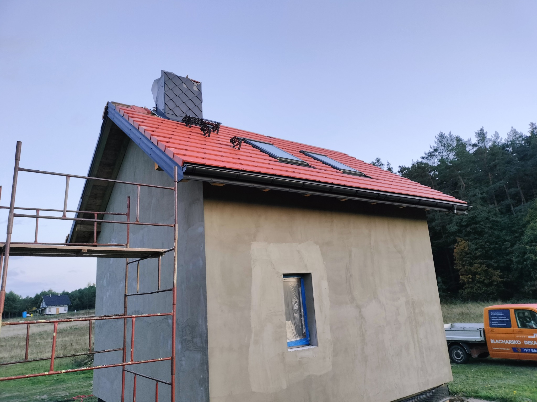 Niedawno ukończony budynek z czerwonym dachem, dwoma oknami dachowymi, kominem z szarej dachówki łupkowej i nowymi rynnami, częściowo zakryty rusztowaniem, na tle zalesionego wzgórza.