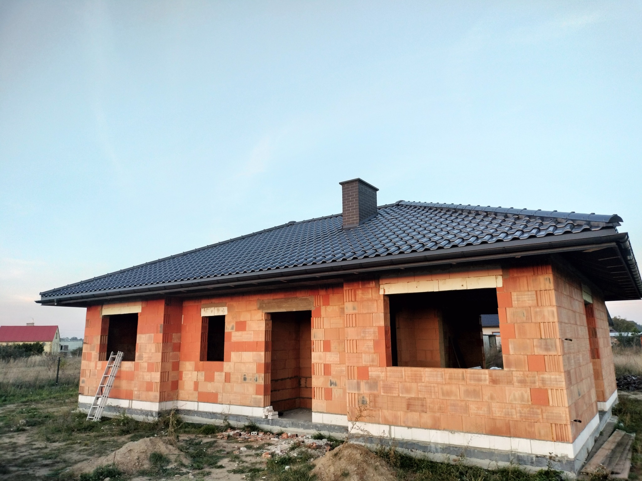 Niedokończony dom jednorodzinny z ciemną dachówką i ceglanym kominem, widoczna drabina oparta o ścianę, stan surowy otwarty.