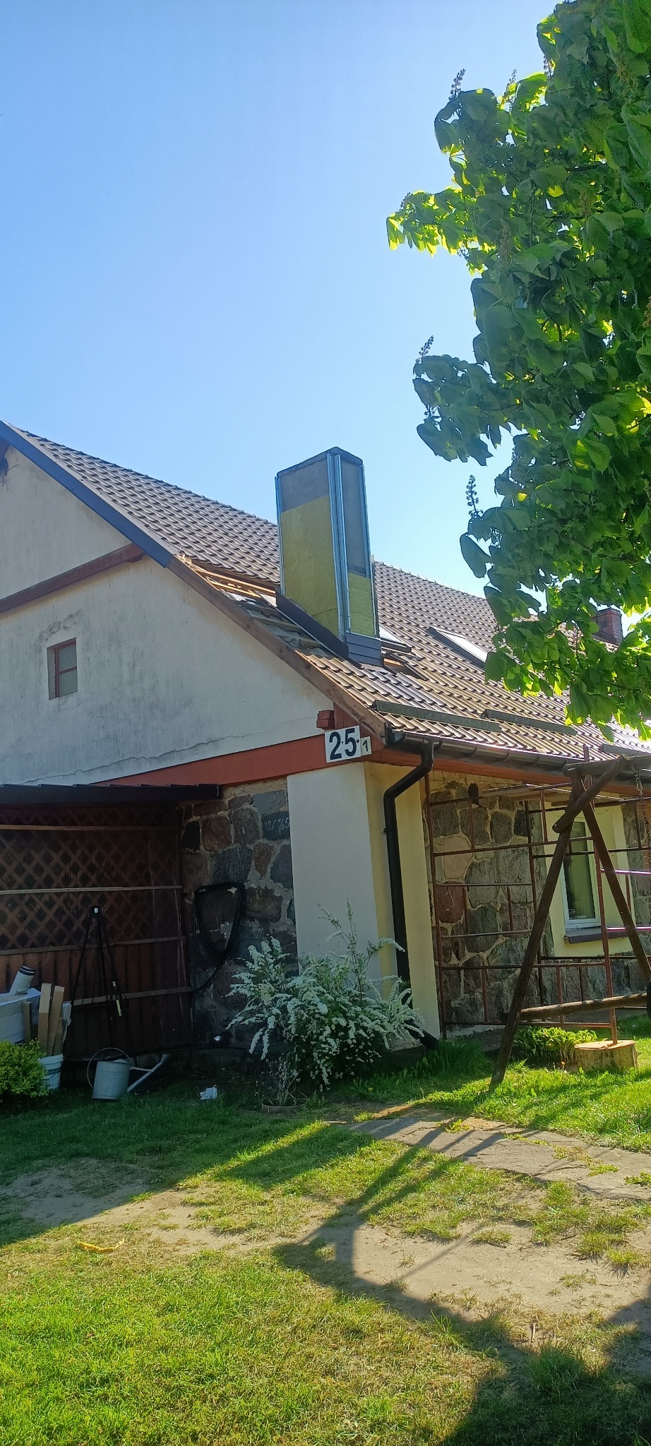 Dom jednorodzinny z brązowym dachem, na którym widoczny jest komin w trakcie montażu, zabezpieczony żółtą izolacją. Część dachu jest odsłonięta, a w tle błękitne niebo i fragment drzewa.