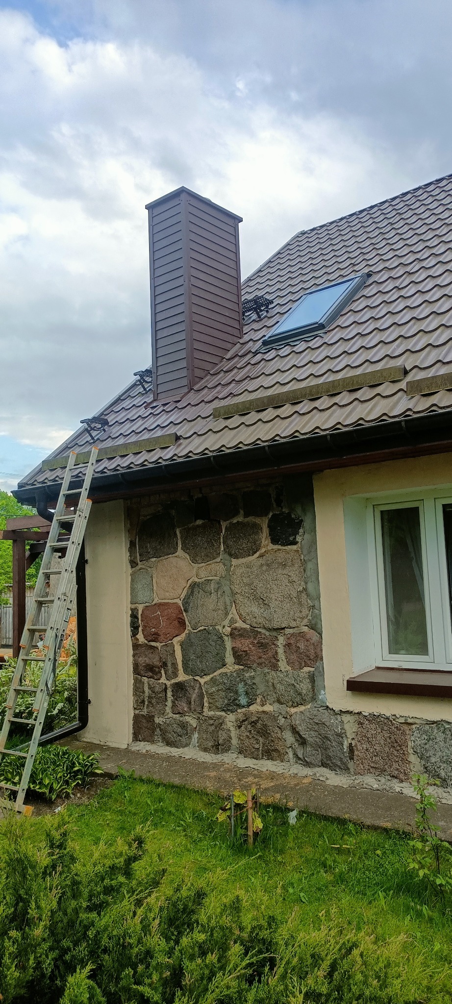 Dom z brązowym dachem z dachówki karpiówki, kominem w kolorze dachu, oknem dachowym i kamienną elewacją. Przystawiona drabina sugeruje prace na wysokości.