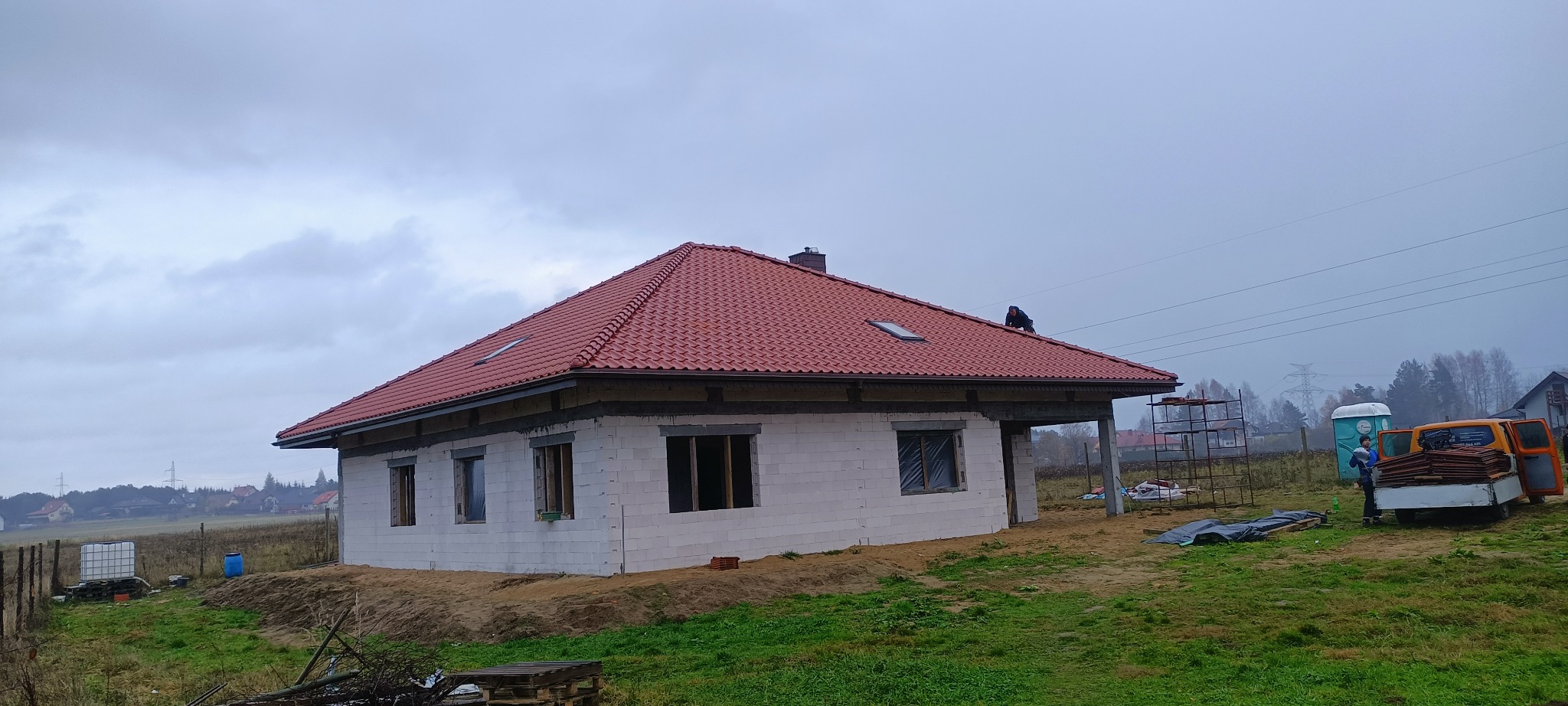 Dom w trakcie budowy z nowym, czerwonym dachem z dachówki ceramicznej; widoczny dekarz na dachu oraz częściowo wykończona elewacja z białego bloczka.