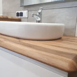 Biała, owalna umywalka nablatowa na blacie z drewna egzotycznego o zaokrąglonych krawędziach w nowoczesnej łazience z szarą płytką ceramiczną.