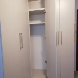 Biała szafa z otwartymi drzwiami, widoczne półki wewnątrz, minimalistyczne uchwyty, szara podłoga.