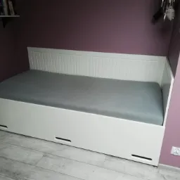 Biała, drewniana sofa z szarą pościelą w pokoju z fioletowymi ścianami, widoczne szuflady pod sofą i półka z dekoracjami w tle.