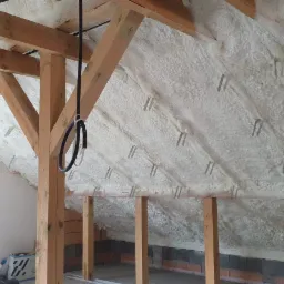 Poddasze z drewnianą konstrukcją krokwiową, pokryte białą, grubą warstwą izolacji natryskowej z widocznymi metalowymi mocowaniami, częściowo otynkowana ściana z lewej strony, fragment muru z cegły...