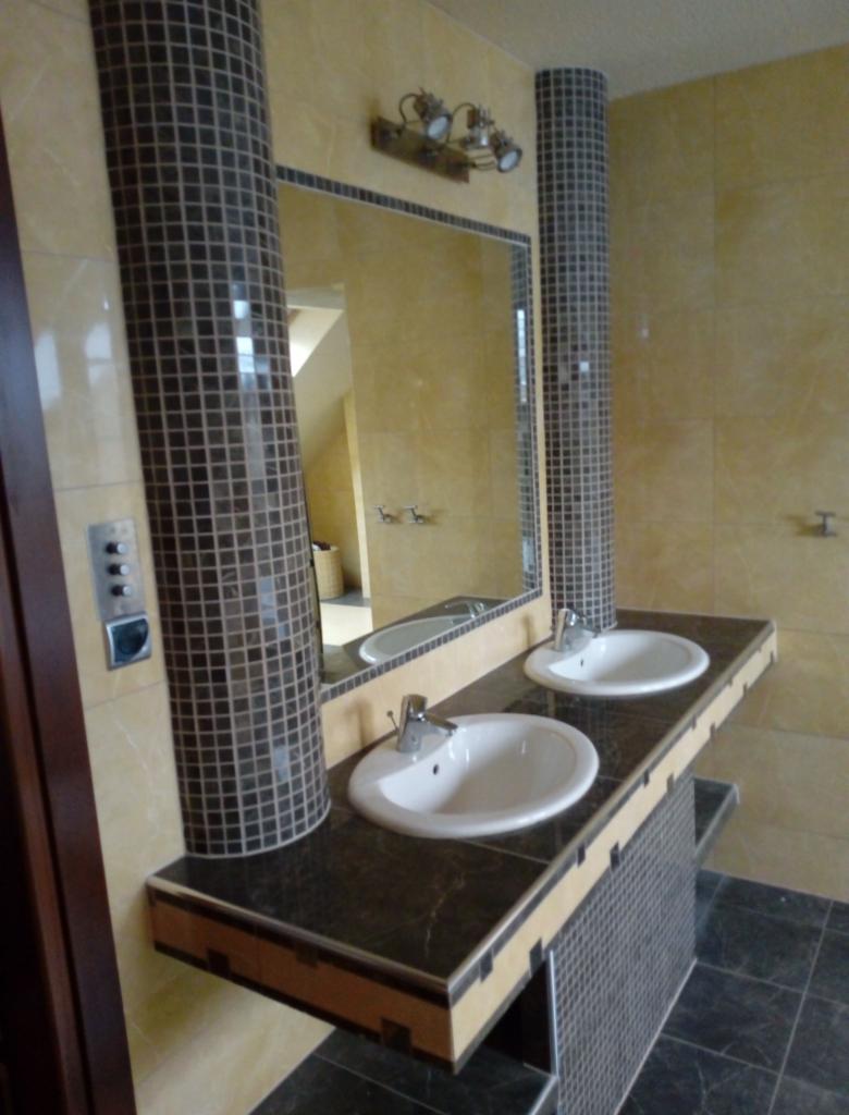 Wykończona łazienka z dwoma białymi umywalkami osadzonymi w ciemnym blacie z mozaikową kolumną po każdej stronie lustra, oświetlenie punktowe na ścianie.