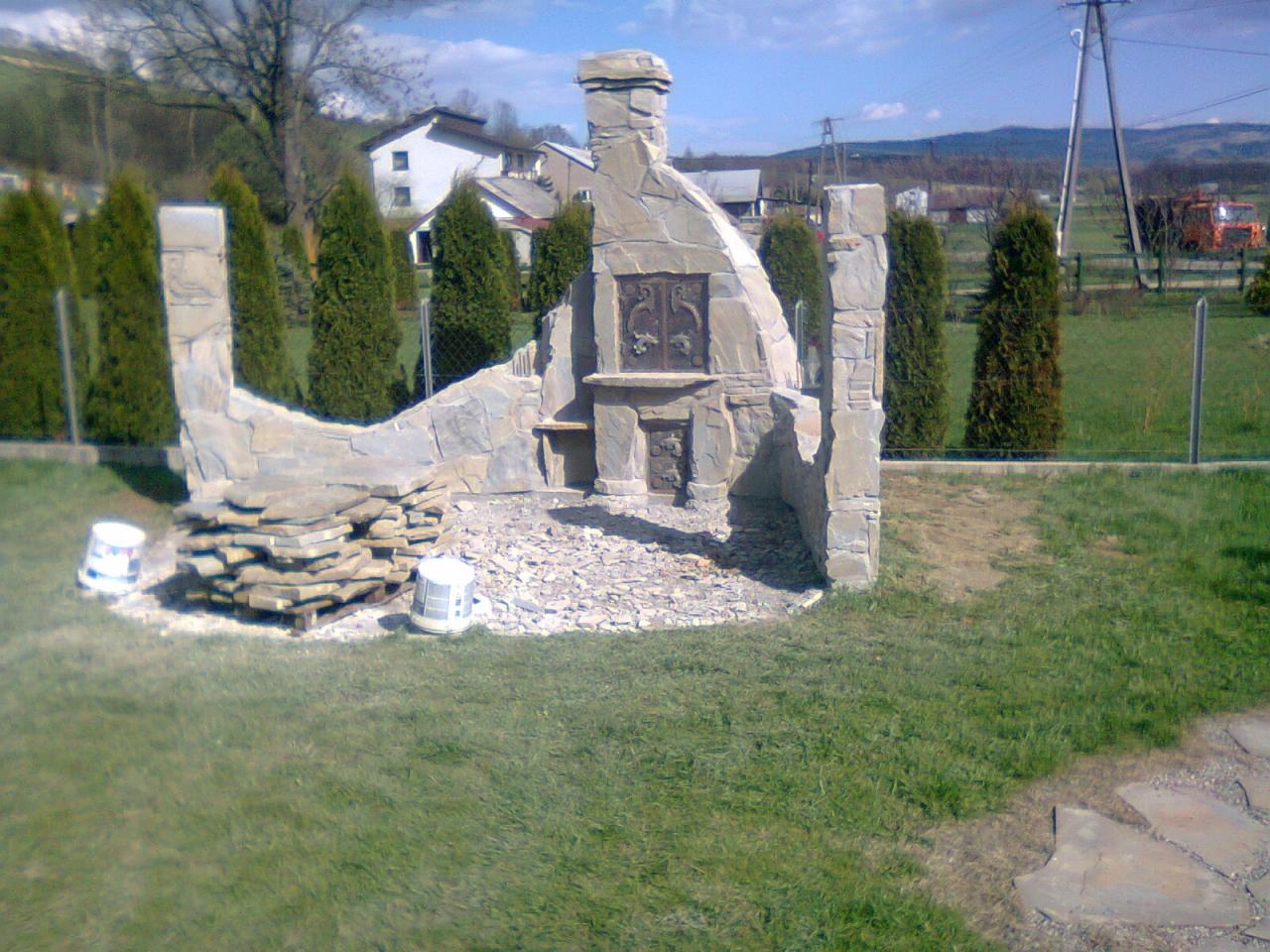 Kamienna konstrukcja grilla ogrodowego w trakcie budowy, z elementami dekoracyjnymi i stosem kamieni obok, otoczona trawnikiem i tujami, w tle zabudowa mieszkalna i wzgórza.