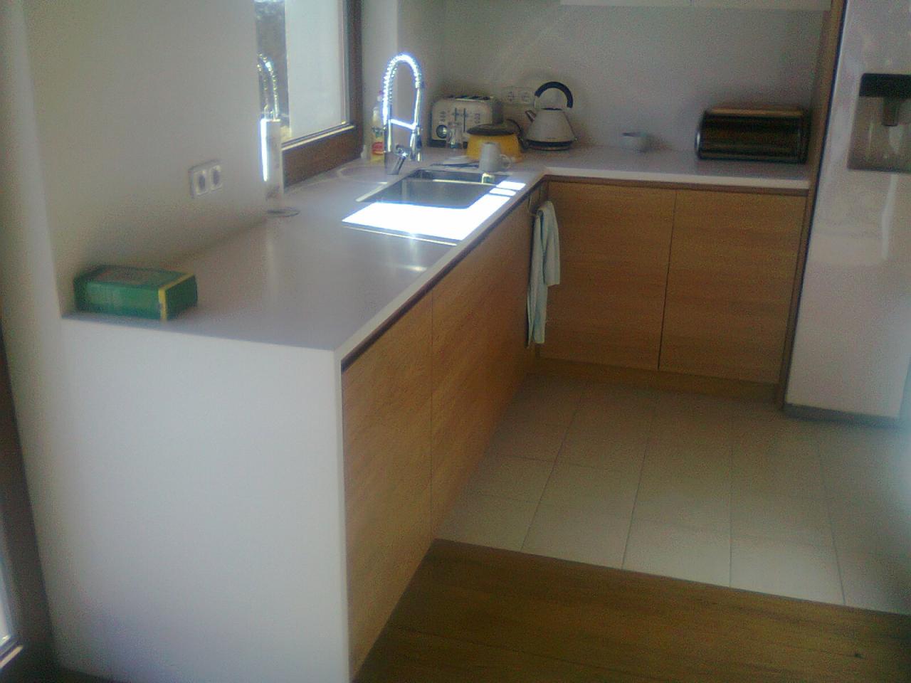 Jasna kuchnia w Bobowej z drewnianymi szafkami, białym blatem i zlewozmywakiem pod oknem, widok na fragment podłogi wyłożonej płytkami.