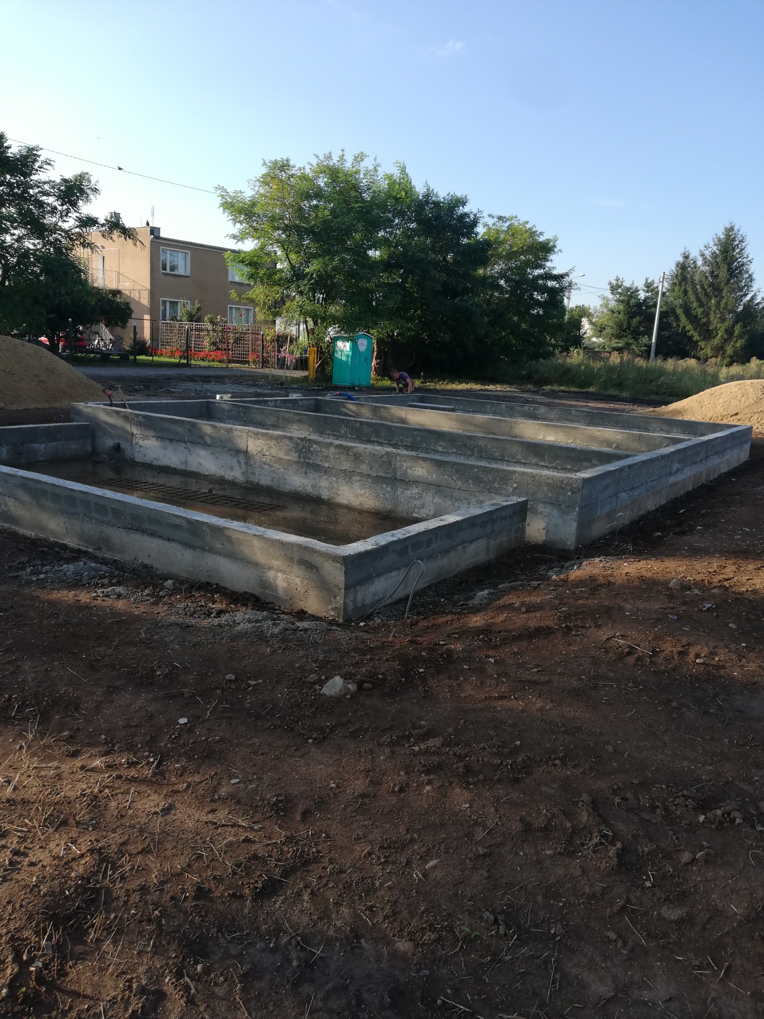 Wylane betonowe ściany fundamentowe z widocznym zbrojeniem wewnątrz wykopu, z hałdą piasku w tle i budynkiem mieszkalnym w oddali.