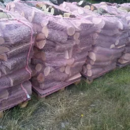 Rząd palet z workami drewna kominkowego, zabezpieczonymi różową siatką, ułożonych na trawie.