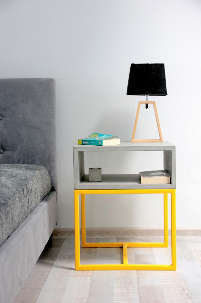 Betonowa szafka nocna z żółtym metalowym stelażem, na niej książka, lampka z czarnym abażurem i betonowa kostka, obok łóżko z szarym zagłówkiem.