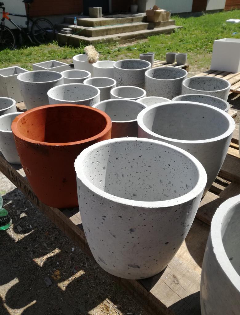 Różnorodne betonowe donice ogrodowe, okrągłe i kwadratowe, w odcieniach szarości i terakoty, ustawione na palecie w słoneczny dzień, z rowerem i schodami w tle.