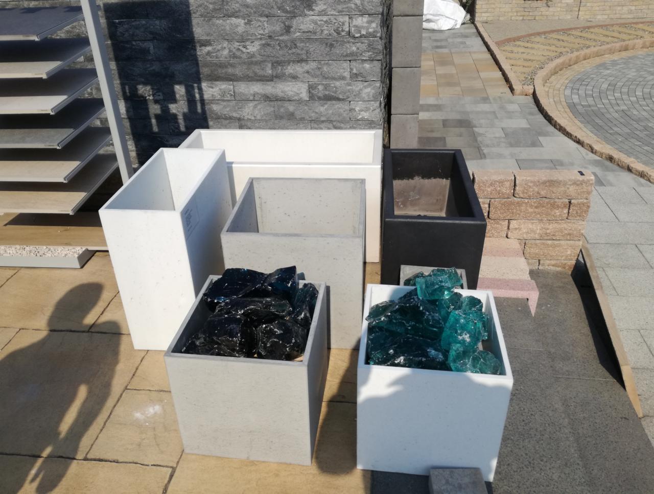 Ekspozycja betonowych donic w różnych kolorach (biały, szary, czarny) wypełnionych dekoracyjnym szkłem w odcieniach czerni i turkusu, na tle kostki brukowej i elementów wykończeniowych.
