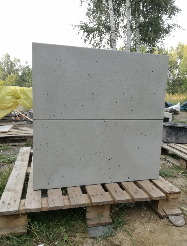 Dwa prefabrykowane, szare elementy betonowe na drewnianej palecie, widoczne na tle zieleni i drzew.