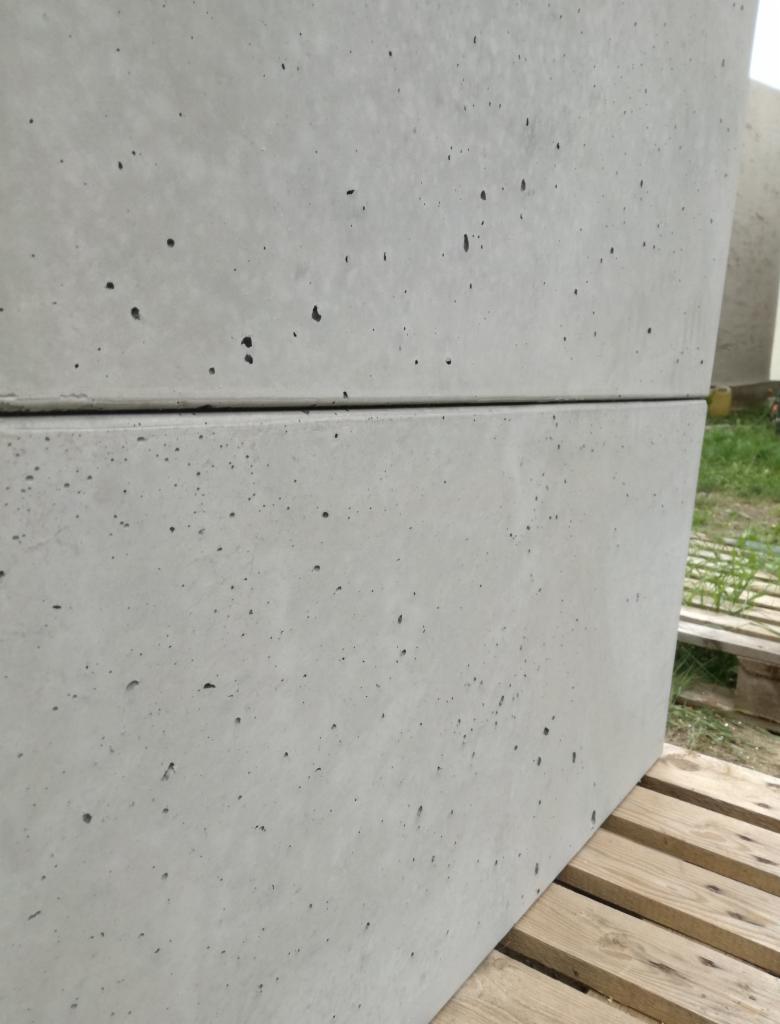 Dwa prefabrykowane elementy betonowe z charakterystycznymi porami powietrznymi, ułożone jeden na drugim, stojące na drewnianej palecie w otoczeniu zieleni.