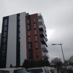 Wielopiętrowy budynek mieszkalny z balkonami, z elewacją w kolorach białym, czarnym i ceglastym, widziany z dołu, z zaparkowanymi samochodami na pierwszym planie i słupem oświetleniowym po prawej...