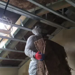 Pracownik w kombinezonie ochronnym i rękawicach montuje izolację z wełny mineralnej na poddaszu, widoczne metalowe profile konstrukcyjne i okno dachowe.