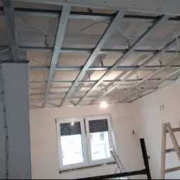 Metalowa konstrukcja sufitu podwieszanego z widocznymi przewodami elektrycznymi, częściowo zakryta płytami kartonowo-gipsowymi, drabina oparta o ścianę, okno z żaluzjami w tle.