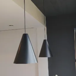 Nowoczesne, stożkowe lampy wiszące w kolorze grafitowym, oświetlające wnętrze z białymi i grafitowymi ścianami, z widocznymi elementami sufitu podwieszanego i dużymi oknami.