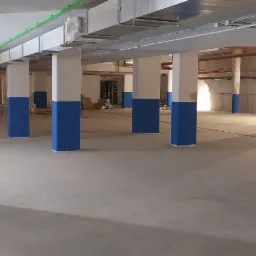 Wnętrze hali produkcyjnej lub magazynowej po malowaniu kolumn na biało i niebiesko, widoczne instalacje pod sufitem i posadzka betonowa.