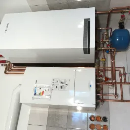 Biały, dwuczęściowy piec grzewczy Biawar z miedzianą instalacją, niebieskim zbiornikiem wyrównawczym i naklejoną etykietą energetyczną na tle szarych płytek