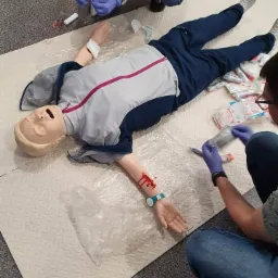 Manekin w sportowym stroju leży na macie, pozorując rannego, podczas gdy dwie osoby w rękawiczkach udzielają mu pomocy, symulując opatrywanie ran.