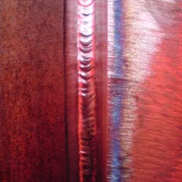 Czerwono-brązowa powierzchnia metalu z wyraźnym, pionowym śladem po spawaniu, widoczne tęczowe przebarwienia termiczne.