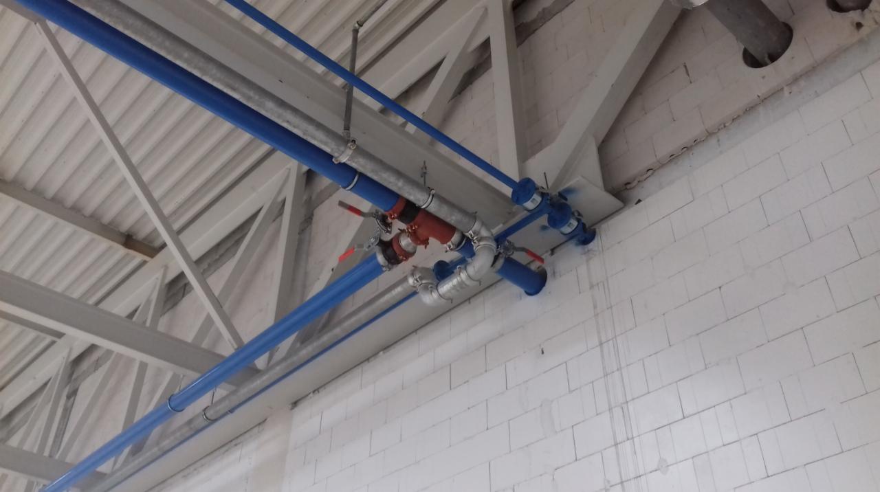 Instalacja przemysłowa z rur w kolorze niebieskim i srebrnym, z zaworami i złączkami, zamontowana pod sufitem na konstrukcji stalowej w hali wykończonej białą cegłą.