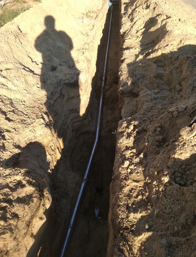 Rów z ułożoną rurą, cień osoby widoczny na skarpie wykopu.