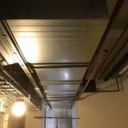 Widok z dołu na system kanałów wentylacyjnych i rur pod sufitem w korytarzu budynku, z widoczną instalacją elektryczną, rurami przeciwpożarowymi i sprzętem do prac na wysokości w tle.