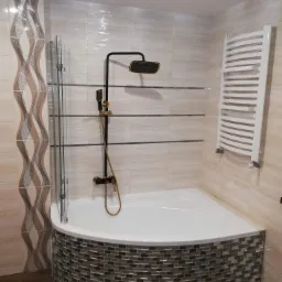 Wykończona łazienka z narożną wanną obudowaną mozaiką, czarna armatura z elementami w kolorze złota, dekoracyjne płytki na ścianie i grzejnik drabinkowy.