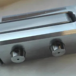 Precyzyjnie obrobiony, metalowy element o złożonym kształcie z widocznymi śladami frezowania i dwoma cylindrycznymi otworami, leżący na jasnym tle.