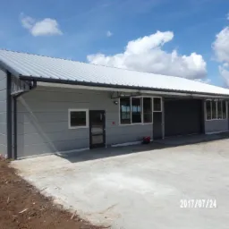 Szary budynek warsztatowy z blachy trapezowej, z bramą rolowaną i oknami, wykończony czarnymi elementami, z datą 2017/07/24 w prawym dolnym rogu.