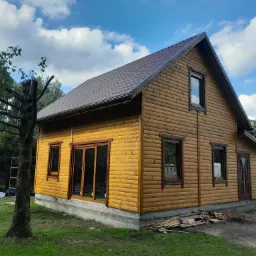 Zewnętrzny widok parterowego domu z bali z brązowym dachem, widoczne okna i drzwi z ciemnymi ramami, częściowo wykończony teren wokół budynku, na trawie leżą resztki drewna, w tle drzewa i błękitne...